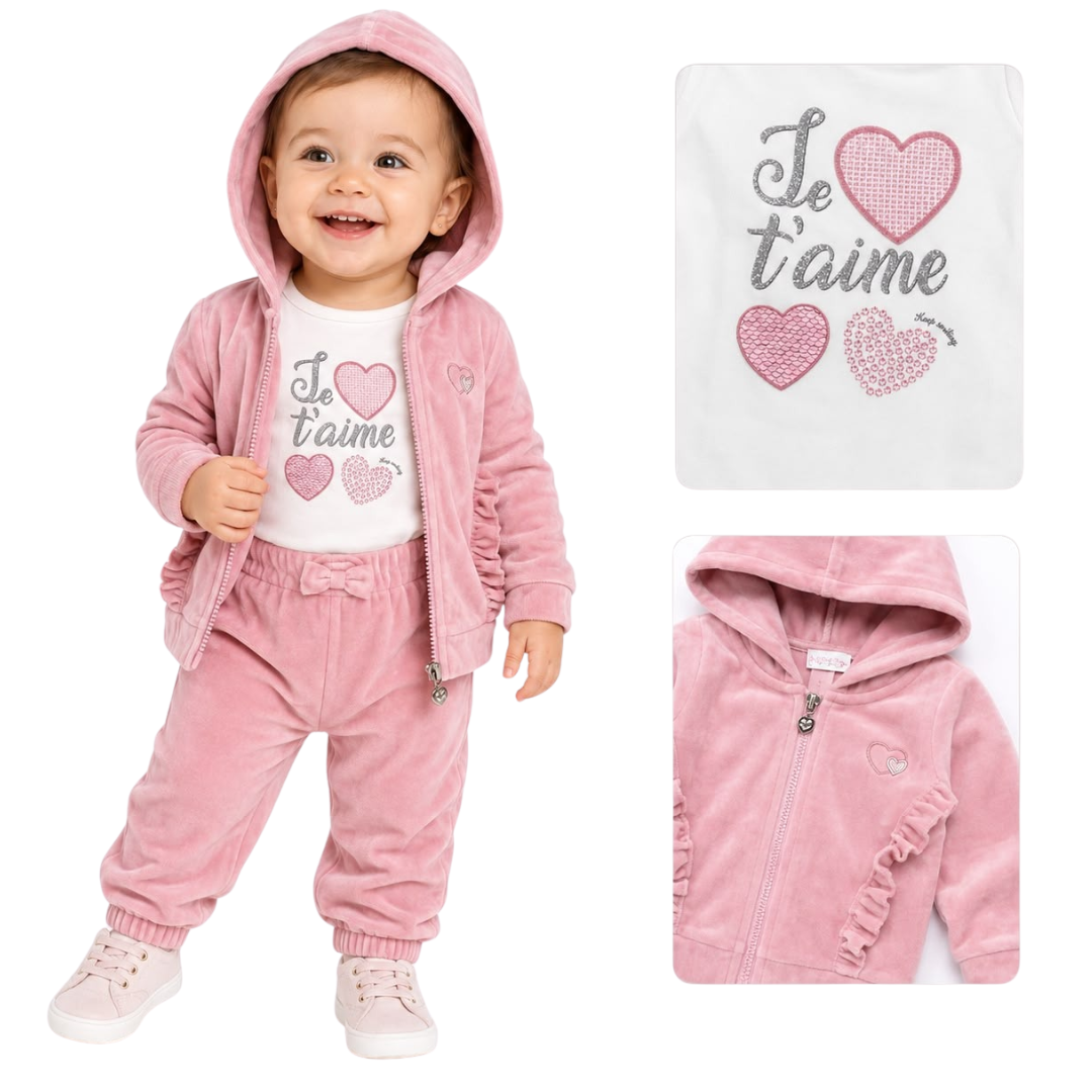 A baby girl wearing a pink hooded zip-up jacket and matching jogger pants over a white graphic t-shirt. The set features a pink velour texture and a white graphic print with pink and gray hearts and the French text 'Je t'aime'.