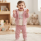 A toddler girl wearing a pink two-piece fleece tracksuit with a zip-up hoodie and matching pants, standing in a bright, cozy room with toys and a teddy bear in the background.