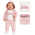 A baby girl wearing a pink two-piece tracksuit with a white t-shirt underneath. The set includes a zip-up hoodie with a hood, matching jogger pants, and a matching square washcloth.