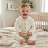 A baby lying on a cream-colored textured bassinet, wearing a light blue and white long-sleeve romper with a blue train graphic on the chest and matching light blue pants.