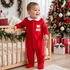Baby wearing a red velour Christmas sleepsuit next to a holiday tree.