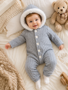 Baby in a gray knitted outfit with white hood lying on a soft surface with a teddy bear in the background.