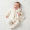 Smiling baby lying on a white surface wearing a cream fleece onesie and cuddling a plush bear security blanket.