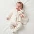 Smiling baby lying on a white surface wearing a cream fleece onesie and cuddling a plush bear security blanket.