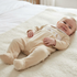 A baby lying on a cream blanket on a bed, wearing a beige footed sleeper with a white chest panel and small embroidered animal details.
