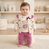 A baby standing on a small beige stool in a nursery, wearing a cream cardigan with teddy bear prints, a matching cream t-shirt with a teddy bear graphic, and magenta pants.