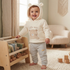 A baby wearing a cream-colored hooded sweatshirt with a teddy bear face and the words 'little one' on the front, paired with light gray jogger pants, standing in a cozy, sunlit nursery next to a wooden toy organizer.
