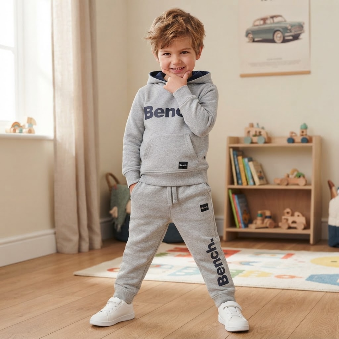 A young boy wearing a light gray two-piece tracksuit with 'Bentley' printed in navy blue on the hoodie and pants, standing in a child's playroom.