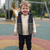 A smiling young boy stands on a colorful playground wearing a dark navy and olive green quilted puffer vest over a cream long-sleeve shirt and black pants.