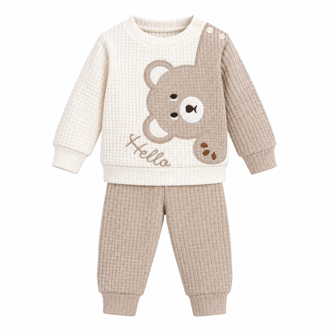 Flat lay product photograph of the Beyaz Bebek cream and beige waffle-knit toddler outfit set against a clean white background, featuring a long-sleeve sweatshirt with an embroidered bear face and the word "Hello," shoulder button closures, and matching ribbed-cuff joggers.