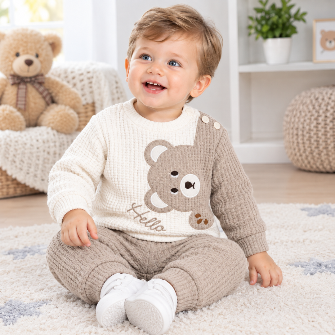 A happy toddler boy sits on a star-patterned rug in a brightly lit nursery, wearing the cream and beige waffle-knit "Hello" bear sweatshirt and matching joggers, with a large teddy bear and cozy room decor softly blurred in the background.
