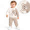 A toddler boy stands smiling against a white background, wearing a cream and beige waffle-knit long-sleeve sweatshirt with a cute embroidered bear patch and the word "Hello," matching waffle joggers, and white sneakers, with a circular inset highlighting a close-up of the bear design and shoulder button detail.