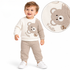 A toddler boy stands smiling against a white background, wearing a cream and beige waffle-knit long-sleeve sweatshirt with a cute embroidered bear patch and the word "Hello," matching waffle joggers, and white sneakers, with a circular inset highlighting a close-up of the bear design and shoulder button detail.
