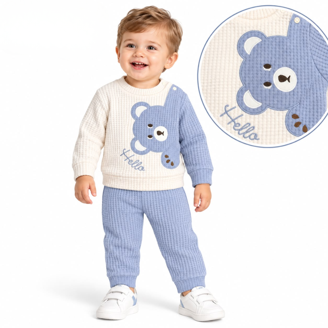 A toddler stands against a white studio background wearing a cream and light blue waffle-knit one-piece outfit with a bear face graphic and the word 'Hello' on the front. A circular inset at the top right shows a close-up of the blue bear face and waffle texture.