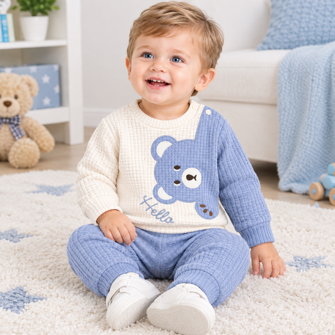 A smiling toddler sitting on a white rug in a cozy room, wearing a waffle-knit two-piece pajama set with a cream top featuring a blue bear graphic and the word 'Hello,' and matching blue pants.