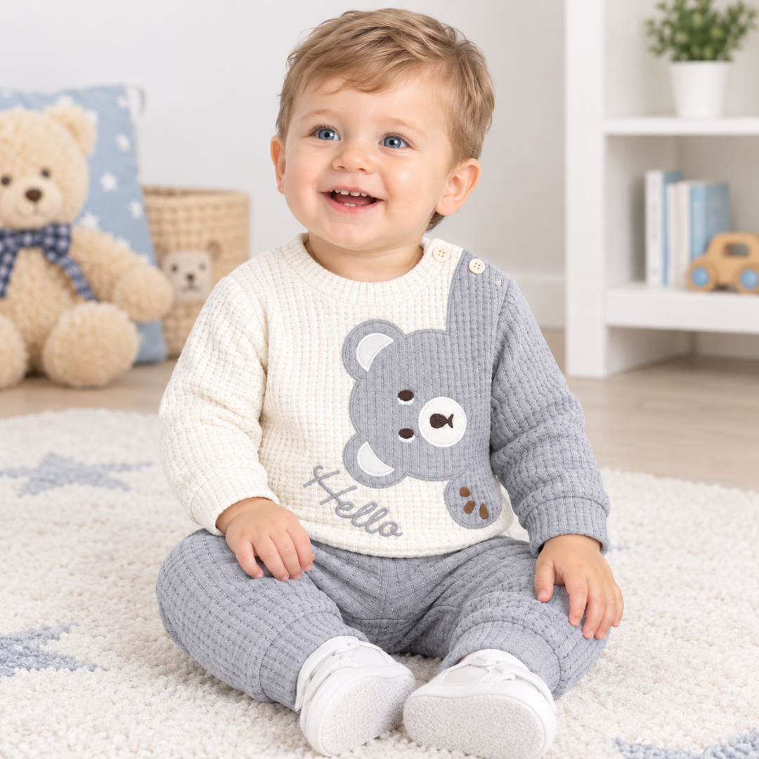 A smiling toddler sits on a white rug in a bright nursery, wearing a waffle-knit two-piece outfit with a cream bib-style top featuring a gray bear face and the word 'Hello,' plus gray pants and white shoes.