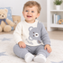 A smiling toddler sits on a white rug in a bright nursery, wearing a waffle-knit two-piece outfit with a cream bib-style top featuring a gray bear face and the word 'Hello,' plus gray pants and white shoes.
