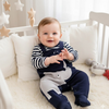 Baby sitting in a crib wearing a striped shirt and navy blue overalls with a white background