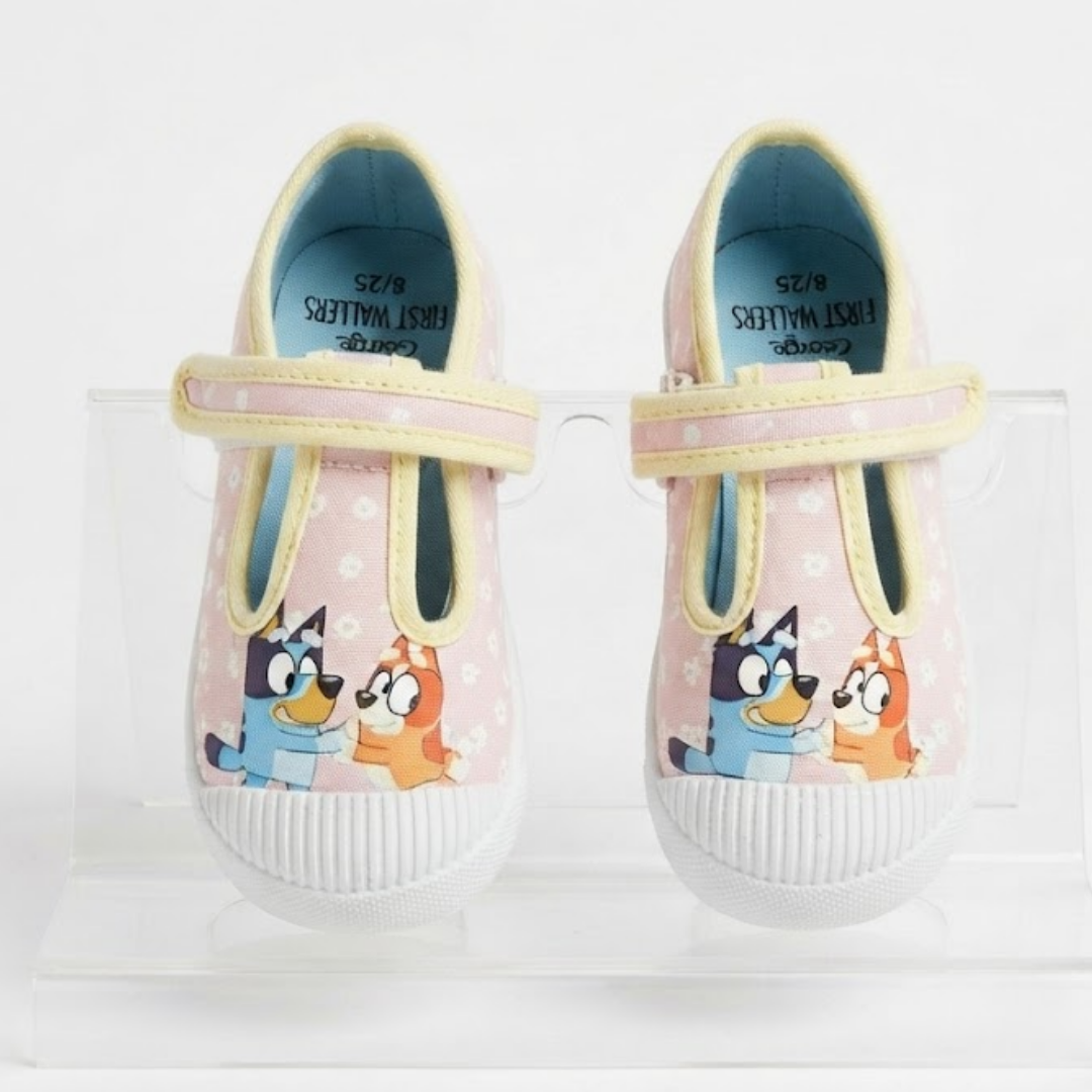 A pair of children's slip-on shoes with a light pink polka-dot upper, featuring a cartoon Bluey character print on the front, a pale yellow strap, and white ribbed soles. They are displayed on a clear acrylic stand against a white background.
