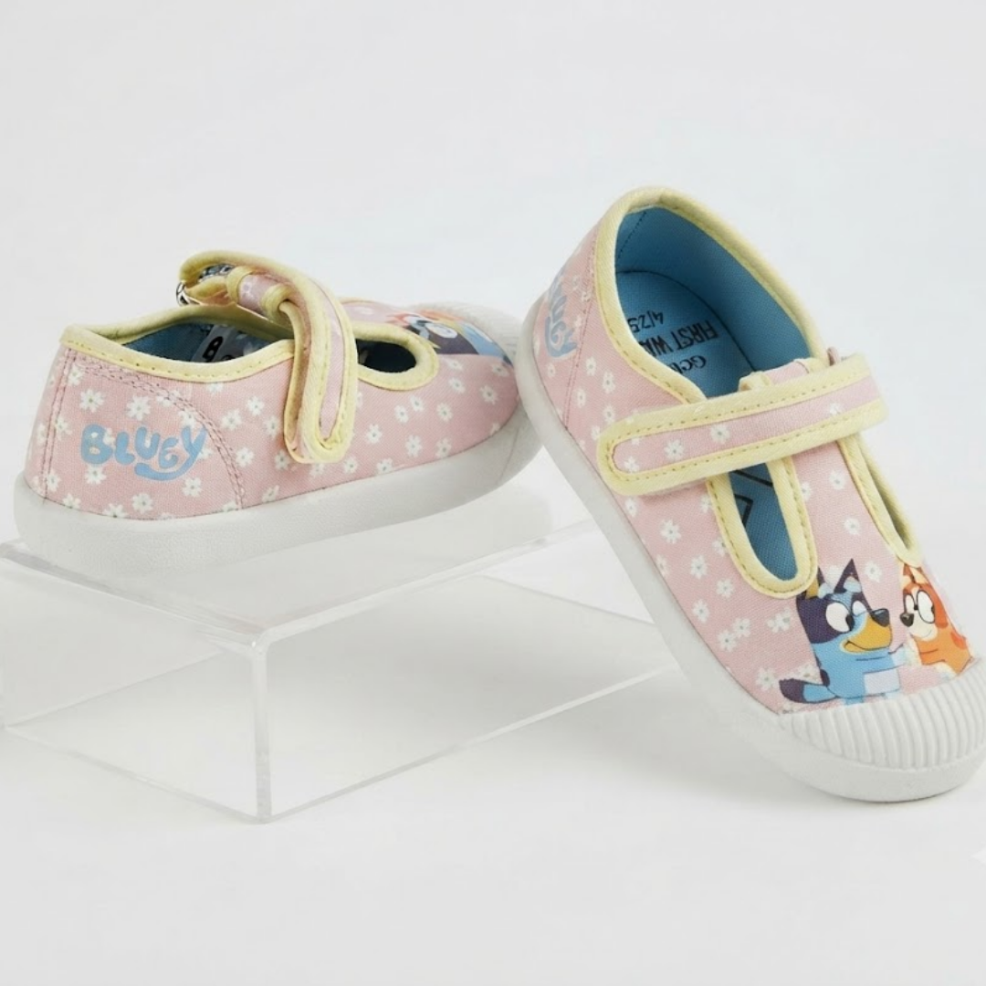 A pair of children's Mary Jane style shoes in light pink with white floral prints, yellow trim, and cartoon character graphics on the toe area. One shoe is displayed on a clear acrylic riser against a white background.