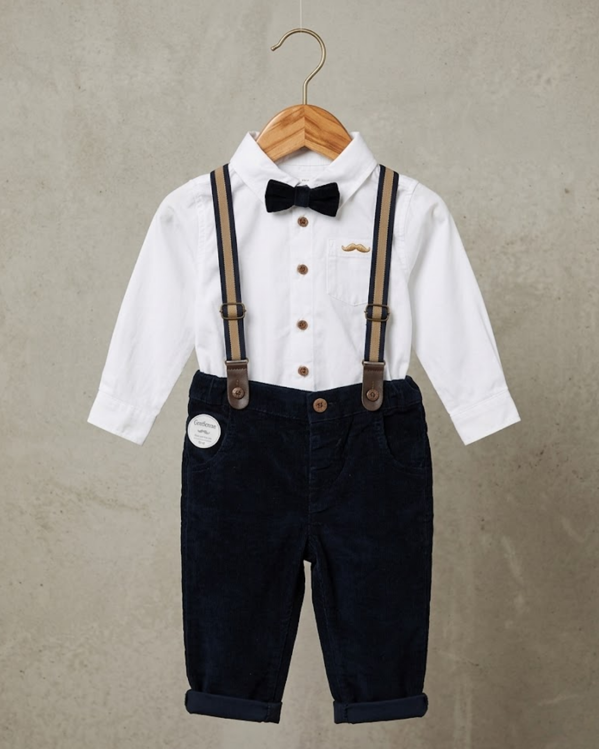Children's outfit with white shirt, black pants, suspenders, and bow tie on a hanger against a beige background.