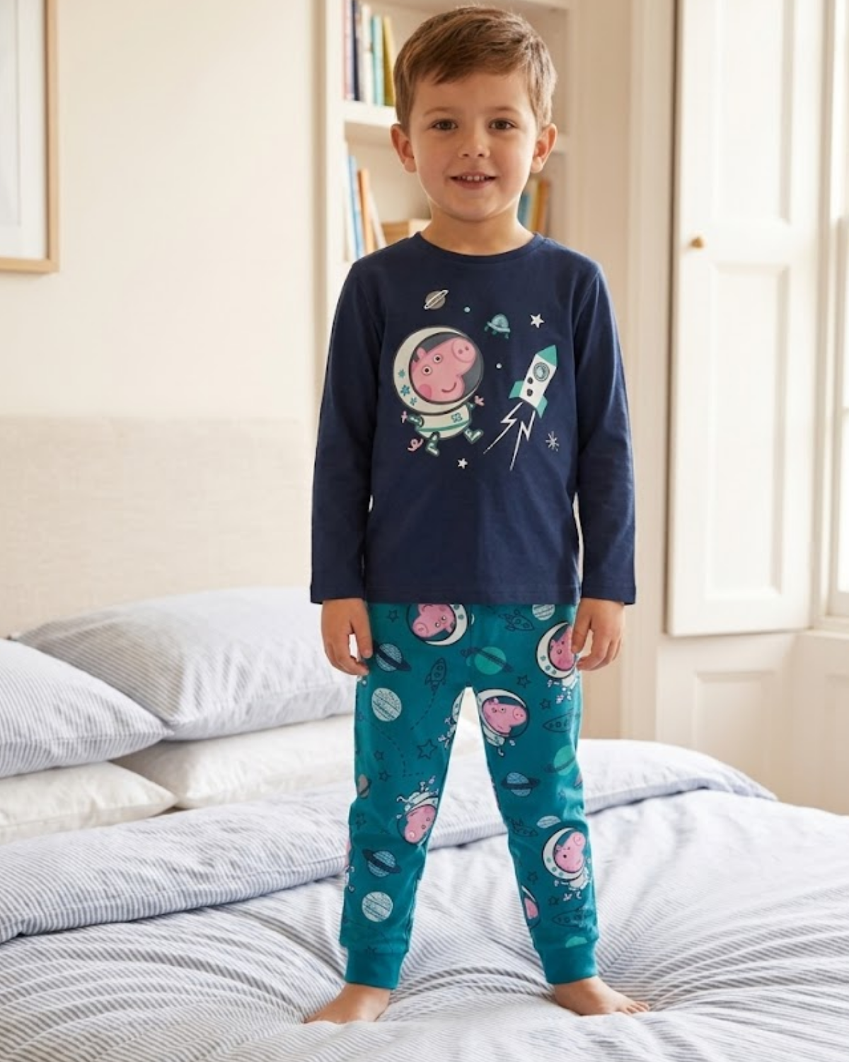 Child wearing space-themed pajamas standing on a bed in a bedroom.