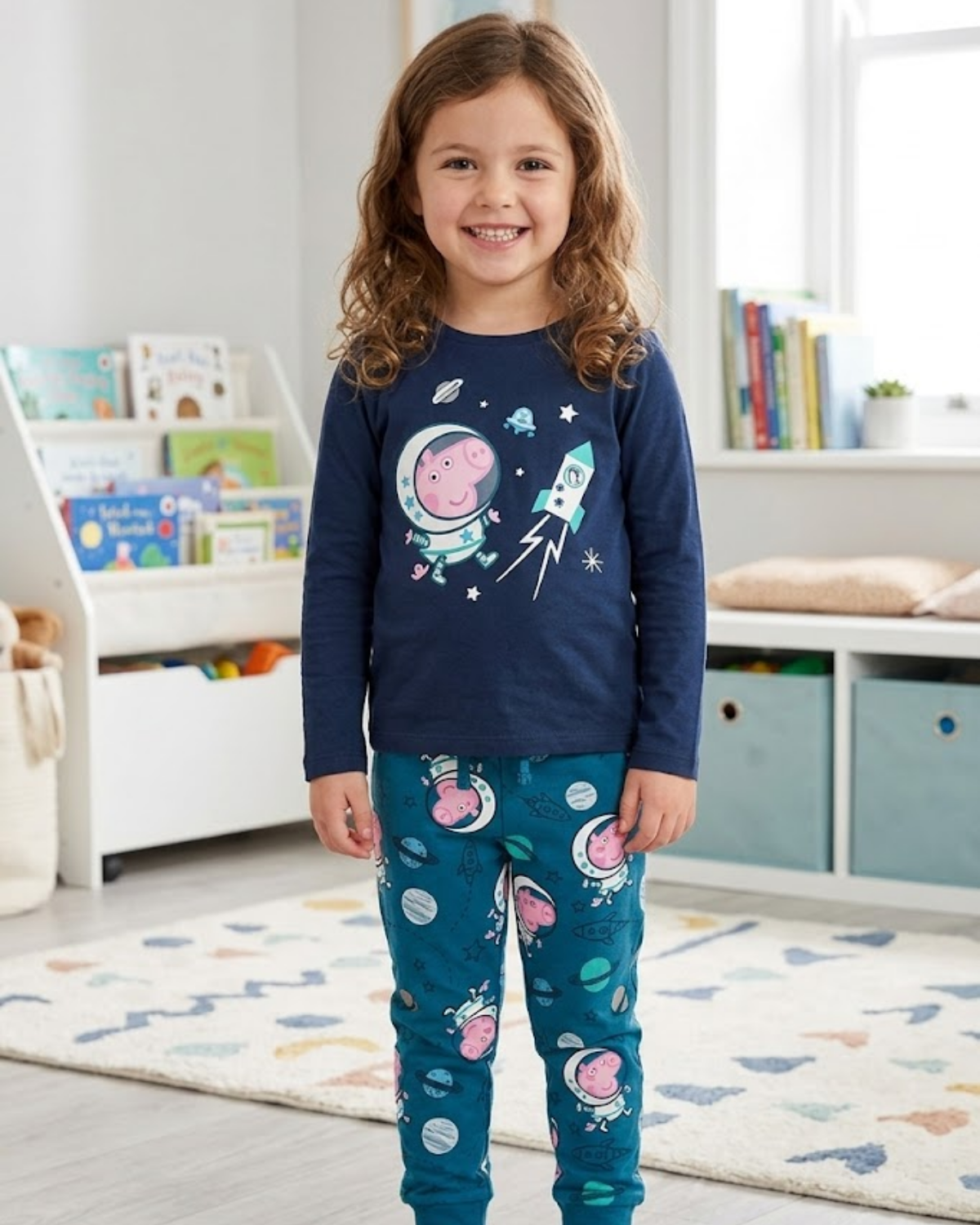 Child wearing a navy blue long-sleeve top and teal pants with space-themed designs in a room with books and toys.
