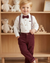 Child in formal attire with suspenders and bow tie standing in front of a wooden shelf with teddy bears and blocks.