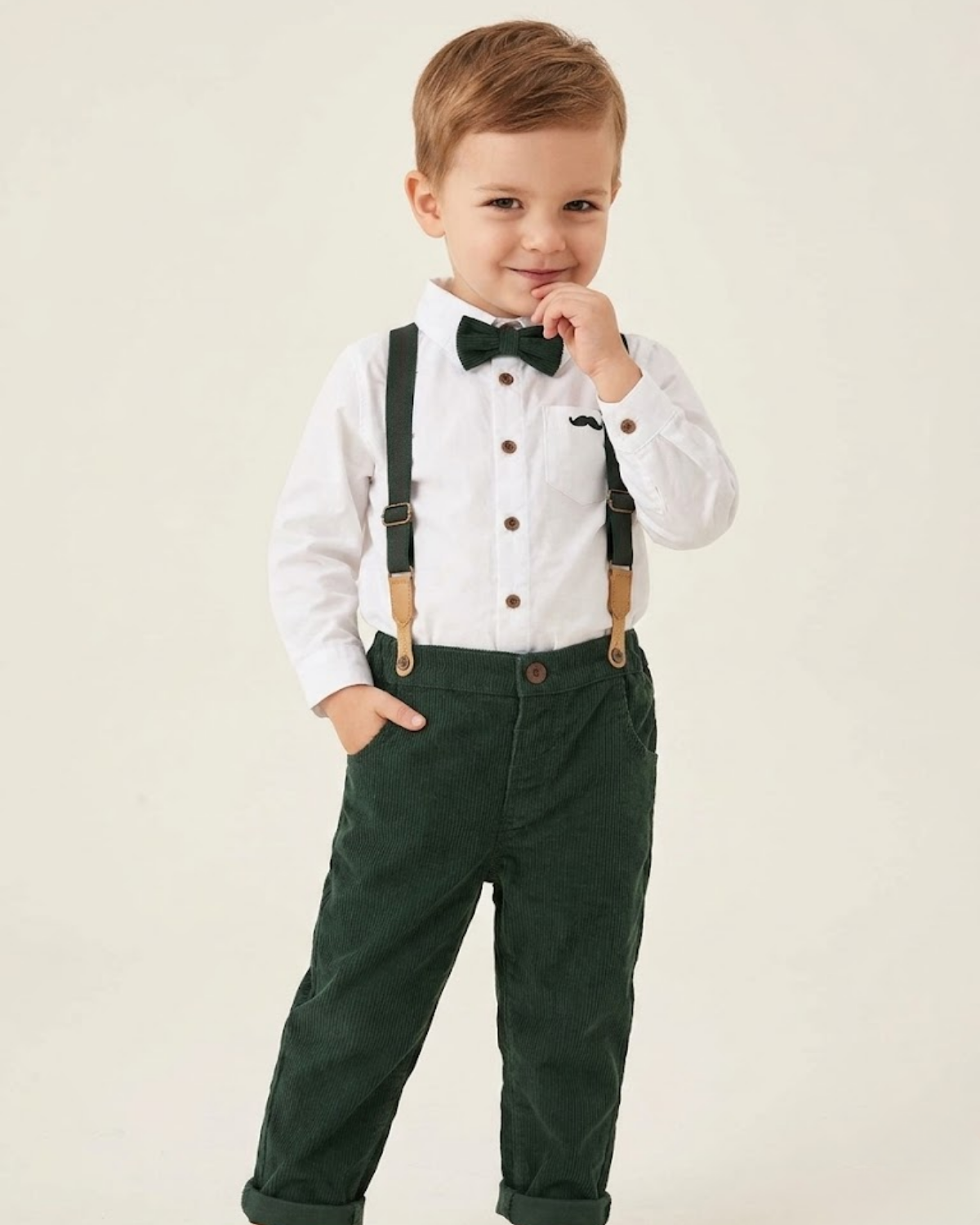 Child wearing a white shirt, green pants, suspenders, and a bow tie on a plain background