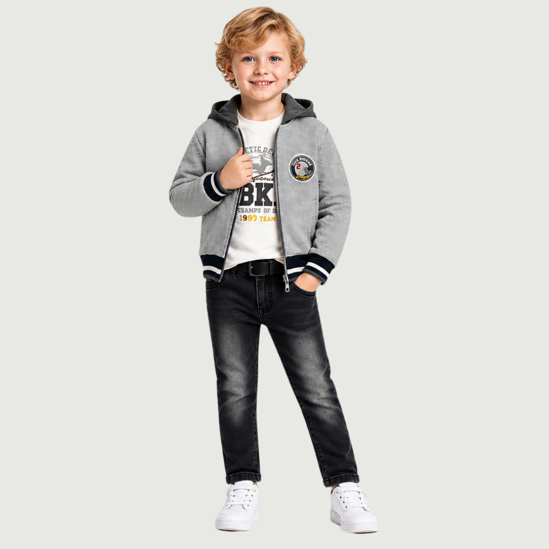 A young boy stands against a light gray background wearing a gray zip-up hooded jacket with black and white striped ribbed cuffs and hem, a white graphic T-shirt, dark gray jeans, and white sneakers.