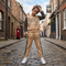 A young boy stands on a cobblestone street wearing a beige two-piece tracksuit with black lace sleeves and side panels, featuring 'Boy' written across the chest and 'BN' on the thigh. He is wearing white sneakers and posing in a town setting with brick buildings in the background.