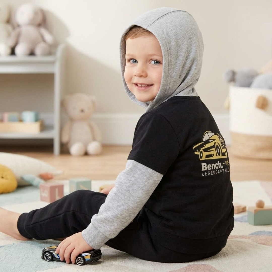 A smiling young child wearing a black and gray hooded sweatshirt sits on a play mat in a cozy, softly lit room with toys in the background.