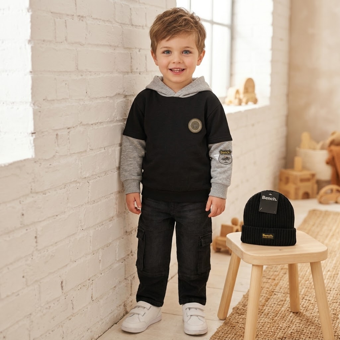 A young boy stands in a bright room wearing a black and gray hooded sweatshirt, dark cargo pants, and white sneakers. A matching black knit beanie with a visible Bench label sits on a small wooden stool beside him.