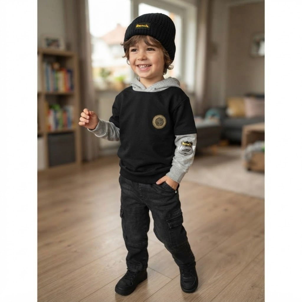 A young child wearing a black and gray hooded sweatshirt with cargo-style pants and a black knit beanie, standing in a softly lit indoor room with wood floors and blurred furniture in the background.