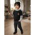 A young child wearing a black and gray hooded sweatshirt with cargo-style pants and a black knit beanie, standing in a softly lit indoor room with wood floors and blurred furniture in the background.