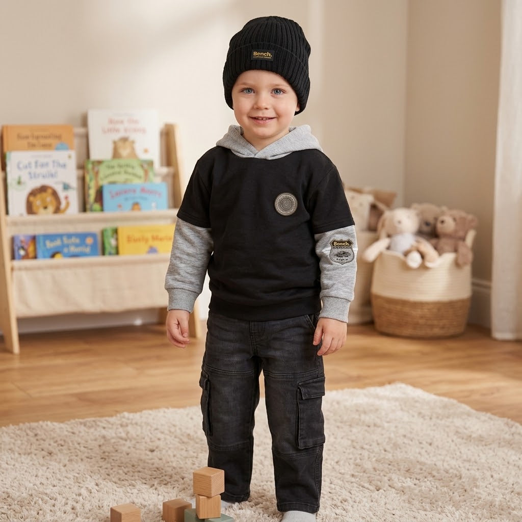 A toddler stands in a child's playroom wearing a black knit beanie with a small gold logo, a black and gray hooded sweatshirt, dark cargo pants, and white socks.