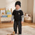 A toddler stands in a child's playroom wearing a black knit beanie with a small gold logo, a black and gray hooded sweatshirt, dark cargo pants, and white socks.