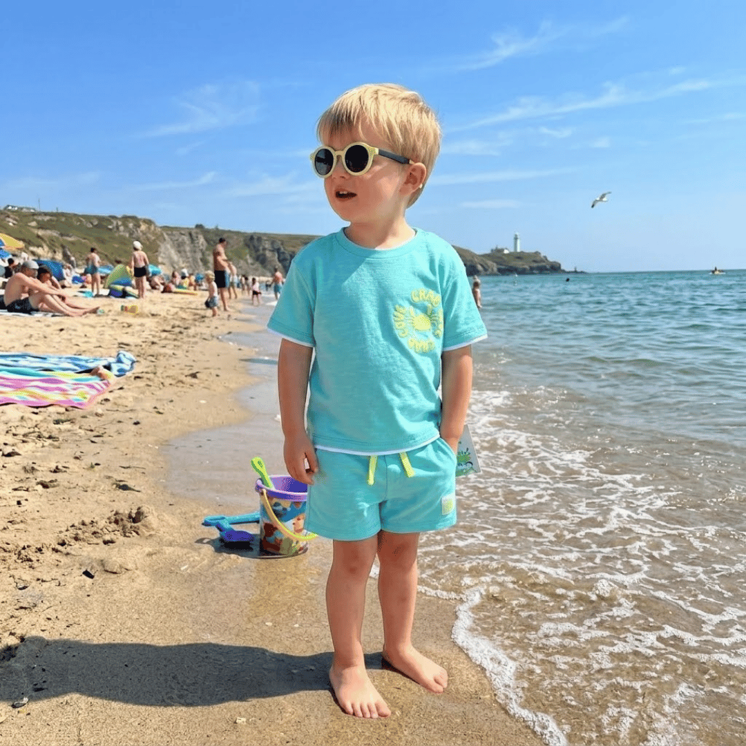 Child in a turquoise shirt and shorts standing on a beach with sunglasses, looking surprised.
