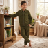 A young boy wearing an olive green two-piece tracksuit with a zip-up jacket and matching pants, standing barefoot in a cozy, sunlit living room.