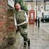 A child wearing an olive green and white 'BOX' sweatshirt and matching sweatpants stands on a wet city sidewalk next to a brick wall, with a red telephone booth and buildings in the background.