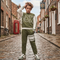 A child stands on a cobblestone street wearing an olive green and white BOX brand sweatshirt and matching joggers with white sneakers.