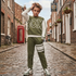 A child stands on a cobblestone street wearing an olive green and white BOX brand sweatshirt and matching joggers with white sneakers.