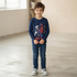 A young boy with tousled brown hair and blue eyes is standing indoors, smiling, wearing a navy long-sleeve t-shirt with a Spider-Man graphic, dark blue jeans, and grey sneakers.
