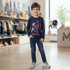 A young boy with tousled brown hair and blue eyes stands centered in a brightly lit children's clothing store, smiling at the camera. He has one hand in his jeans pocket and is wearing a navy long-sleeve t-shirt with a Spider-Man graphic, dark blue jeans, and grey-and-white sneakers. Behind him, store shelves display various children's apparel, toys, and wooden blocks, with a large 'M' logo visible on a counter to the right