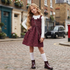 A young girl stands on a cobblestone street wearing a burgundy floral pinafore dress over a white long-sleeve top, with white knee-high socks and dark brown rain boots. A small chain-strap bag is also visible.