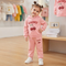A young girl wearing a pink two-piece tracksuit with a cherry graphic on the sweatshirt and pants, standing in a children's clothing store.