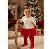 A baby toddler girl wearing a Christmas-themed outfit consisting of a cream long-sleeve top with a reindeer graphic, a red glittery tiered tutu skirt, and red booties, standing in a cozy holiday living room next to a wooden chair.