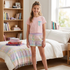 A young girl stands in a bright bedroom wearing a light pink short-sleeve pajama top with a small blue pocket and pastel plaid pajama shorts. The room includes a bed with white bedding, a wooden bookshelf, and a window.