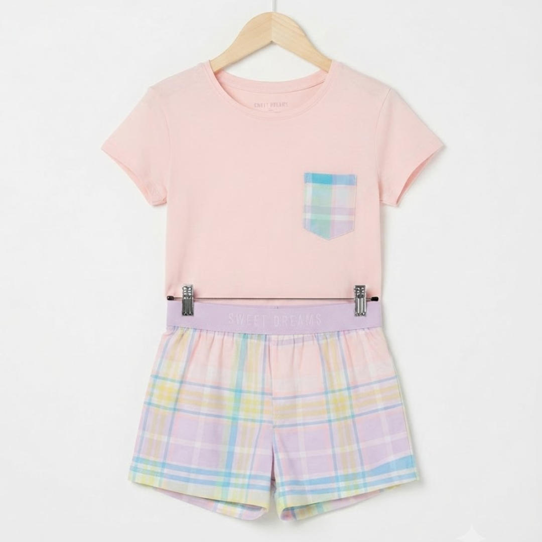 A children's two-piece outfit hanging on a wooden hanger against a plain white background. The set includes a light pink short-sleeve T-shirt with a small plaid pocket on the chest and matching plaid shorts in pastel shades of pink, yellow, blue, and green.