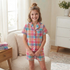 A young girl stands in a cozy bedroom setting wearing a pastel plaid pajama set consisting of a short-sleeve button-up shirt and matching shorts. The pajamas feature a soft checkered pattern in pink, blue, yellow, and white.